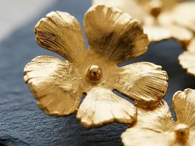 AI-generated macro close-up of the same gold flower earrings on dark slate, brushed metal texture sharp and evenly lit