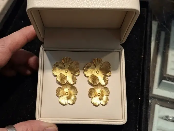 Raw phone photo of gold flower drop earrings in a jewelry store box, held in hand, cluttered counter in the background