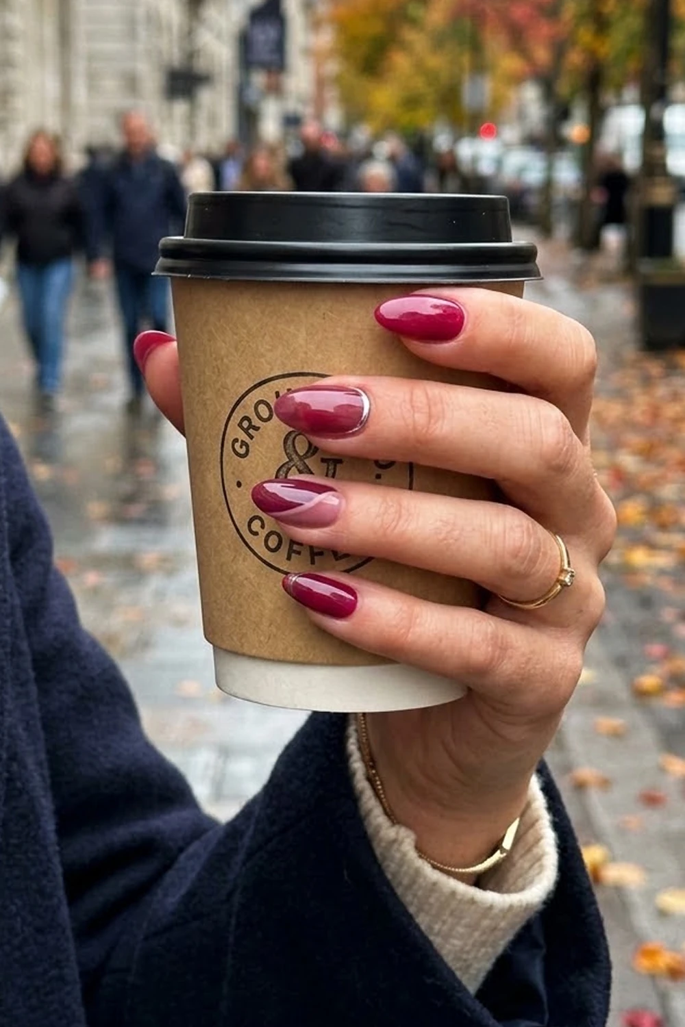 Burgundy glazed almond nails — trending Fall Nails 2026 design shown on a hand holding a coffee cup on an autumn street